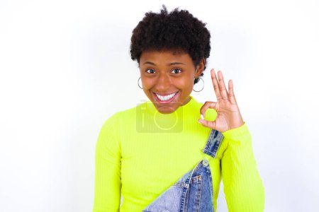Photo for Young African American woman with short hair wearing denim overall against white wall hold hand arm okey symbol toothy approve advising novelty news - Royalty Free Image