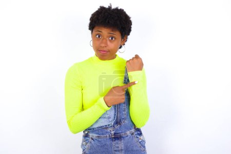 Foto de Joven mujer afroamericana con el pelo corto con denim en general contra la pared blanca en la prisa señalando para ver el tiempo, la impaciencia, molesto y enojado por retraso plazo - Imagen libre de derechos