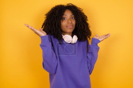Téléchargez les photos : Portrait de jeune femme afro-américaine expressive avec écouteurs sur fond jaune déchiquetant les épaules - en image libre de droit