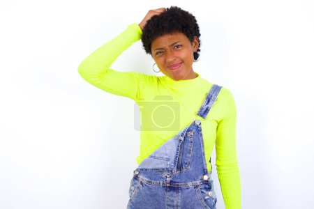 Photo for Young African American woman with short hair wearing denim overall against white wall saying: Oops, what did I do? Holding hand on head with frightened and regret expression. - Royalty Free Image
