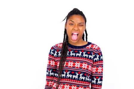 Téléchargez les photos : Femme afro-américaine portant un pull de Noël contre un mur blanc avec un visage heureux et drôle souriant et montrant la langue. - en image libre de droit