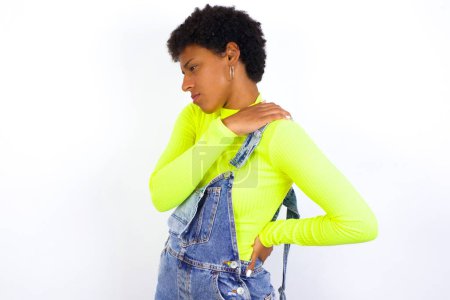 Foto de Joven mujer afroamericana con pelo corto con denim en general contra la pared blanca tiene dolor de espalda - Imagen libre de derechos