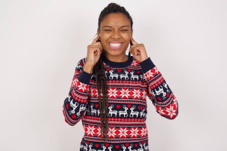 Photo for Stop making this annoying sound! Unhappy stressed out African American woman wearing Christmas sweater against white wall making worry face, plugging ears with fingers, irritated with loud noise. - Royalty Free Image