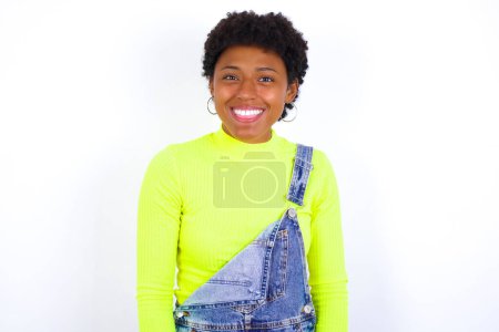 Foto de Joven mujer afroamericana con el pelo corto con denim en general contra la pared blanca con la cara feliz y divertida sonriendo y mostrando la lengua. - Imagen libre de derechos