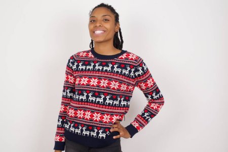 Foto de Estudio de disparo de alegre mujer afroamericana con suéter de Navidad contra la pared blanca mantiene la mano en la cadera, sonríe ampliamente. - Imagen libre de derechos