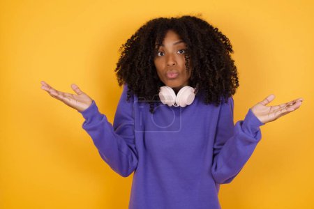 portrait of young expressive african american woman with headphones on yellow background shredding shoulders