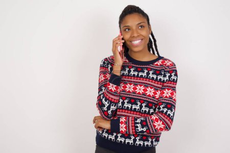 Téléchargez les photos : Portrait d'une femme afro-américaine souriante portant un pull de Noël contre un mur blanc parlant sur un téléphone portable. Concept d'entreprise, confiance et communication. - en image libre de droit