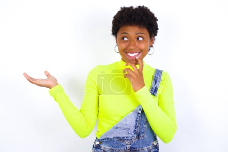 Foto de Positivo joven afroamericana mujer con el pelo corto usando denim general contra la pared blanca anuncio promo toque los dientes de los dedos - Imagen libre de derechos