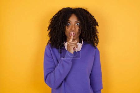 Téléchargez les photos : Portrait d'une jeune femme afro-américaine expressive avec écouteurs montrant un signe de silence sur fond jaune - en image libre de droit