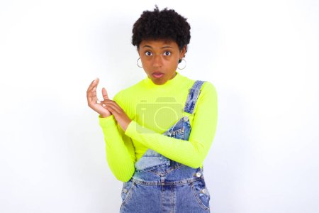 Téléchargez les photos : Surprise émotionnelle jeune femme afro-américaine aux cheveux courts portant le denim dans l'ensemble contre le mur blanc frotte les paumes et regarde la caméra avec incrédulité - en image libre de droit