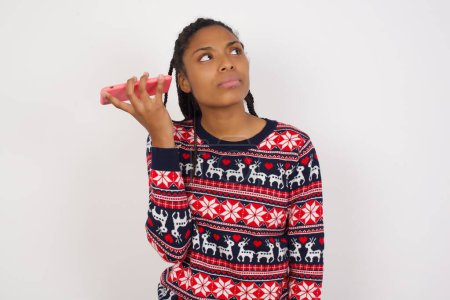 Téléchargez les photos : Femme afro-américaine souriante portant un pull de Noël contre un mur blanc écoutant un message vocal de son smartphone. Concept de communication et technologie. - en image libre de droit