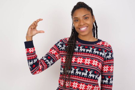 Foto de Mujer afroamericana vistiendo suéter de Navidad contra la pared blanca sonriendo y haciendo gestos con la mano de tamaño pequeño, símbolo de medida. - Imagen libre de derechos