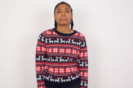 Photo for Gloomy, bored African American woman wearing Christmas sweater against white wall frowns face looking up, being upset with so much talking hands down, feels tired and wants to leave. - Royalty Free Image