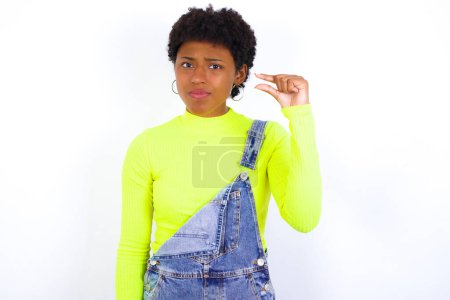 Photo for Upset young African American woman with short hair wearing denim overall against white shapes little gesture with hand demonstrates something very tiny small size. Not very much - Royalty Free Image