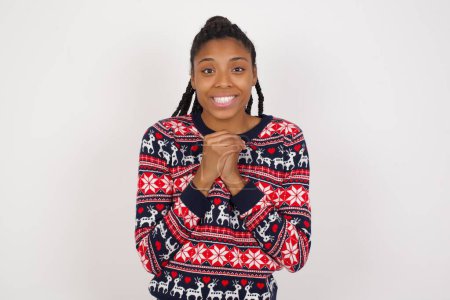 Foto de Mujer afroamericana feliz vistiendo suéter de Navidad contra la pared blanca se levanta contra la pared del estudio naranja mantiene las manos en el corazón, jura ser leal, expresa gratitud. Concepto de honestidad. - Imagen libre de derechos