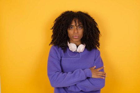 Photo for Portrait of young expressive african american woman with headphones on yellow background with crossed arms - Royalty Free Image