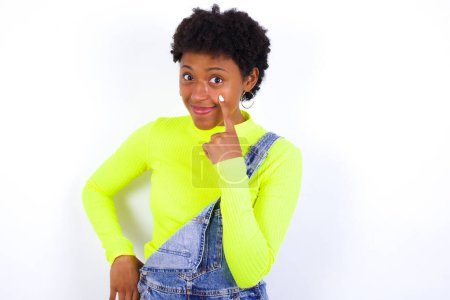 Foto de Mujer afroamericana joven con el pelo corto que usa denim en general contra la pared blanca mirando, observando, manteniendo un ojo en un objeto en frente, o mirando hacia fuera para algo. - Imagen libre de derechos