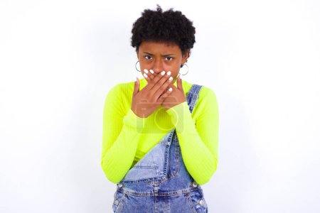 Photo for Upset young African American woman with short hair wearing denim overall against white wall covering her mouth with both palms to prevent screaming sound, after seeing or hearing something bad. - Royalty Free Image