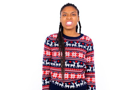 Photo for Mad crazy African American woman wearing Christmas sweater against white wall clenches teeth angrily, being annoyed with coming noise. Negative feeling concept. - Royalty Free Image
