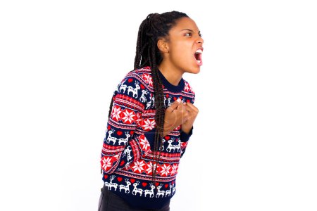 Foto de Mujer afroamericana vistiendo suéter de Navidad contra la pared blanca emocionada y contenta de lograr la victoria, aprieta los puños, grita de emoción con los ojos cerrados, persona exitosa. - Imagen libre de derechos