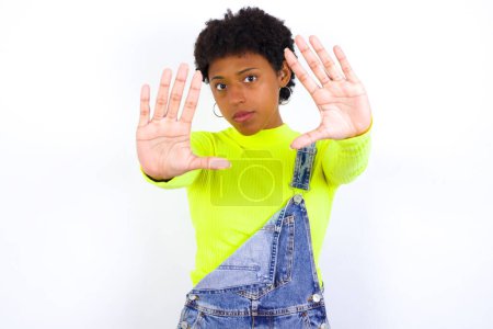 Téléchargez les photos : Portrait de jeune femme afro-américaine souriante aux cheveux courts portant globalement du denim contre un mur blanc regardant la caméra et le cadre gestuel du doigt. Concept de créativité et de photographie. - en image libre de droit