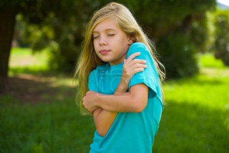 portrait of beautiful caucasian little kid girl wearing blue t-shirt standing outdoor in the park feeling cold