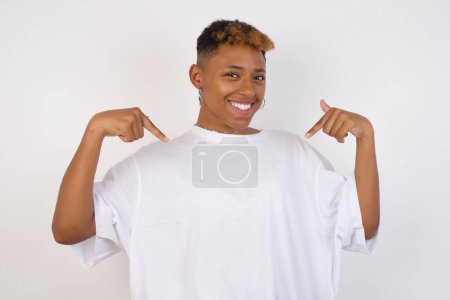Téléchargez les photos : Charmante femme afro-américaine émotive portant un t-shirt blanc pointant vers le bas ou son t-shirt tout en souriant joyeusement et en exprimant des émotions positives sur fond blanc. Fille entraînée beaucoup, elle veut montrer ses muscles - en image libre de droit