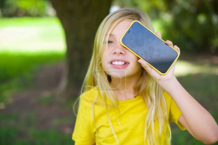 Foto de Niña caucásica vistiendo camiseta amarilla de pie al aire libre sosteniendo teléfono inteligente moderno cubriendo un ojo mientras sonríe - Imagen libre de derechos