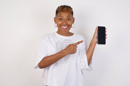 Téléchargez les photos : Femme afro-américaine excitée portant un t-shirt blanc pointant du doigt l'appareil numérique. Studio shot de fille choquée tenant smartphone avec écran vierge. - en image libre de droit