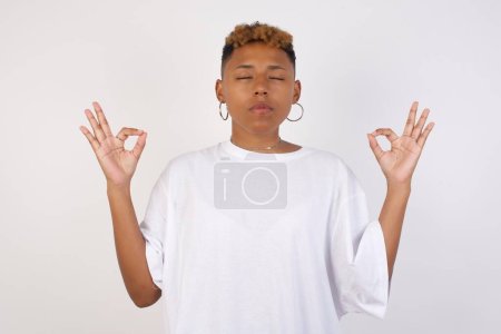 Foto de Meditación, religión y prácticas espirituales. Hermosa joven afroamericana vistiendo camiseta blanca haciendo yoga en la mañana al aire libre después de despertarse, manteniendo los ojos cerrados, sosteniendo los dedos en gesto mudra. - Imagen libre de derechos