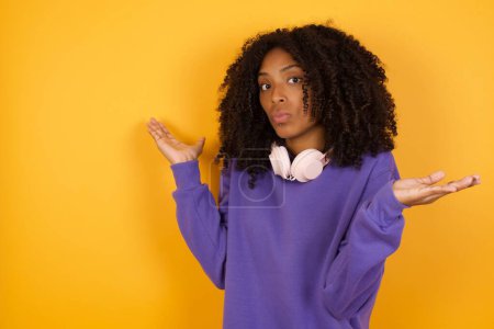 Téléchargez les photos : Portrait de jeune femme afro-américaine expressive avec écouteurs sur fond jaune déchiquetant les épaules - en image libre de droit
