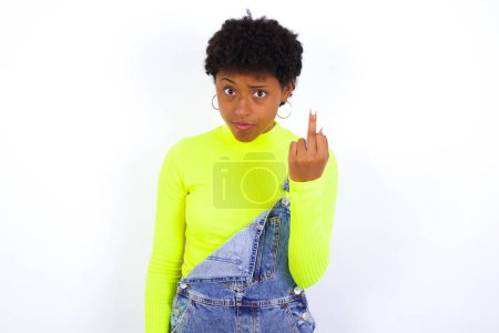 Photo for Young African American woman with short hair wearing denim overall against white wall shows middle finger bad sign asks not to bother. Provocation and rude attitude. - Royalty Free Image