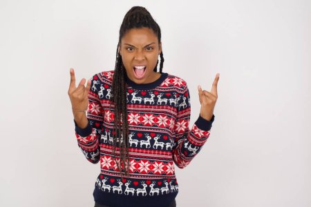Photo for Born to rock this world. Joyful African American woman wearing Christmas sweater against white wall screaming out loud and showing with raised arms horns or rock gesture. - Royalty Free Image