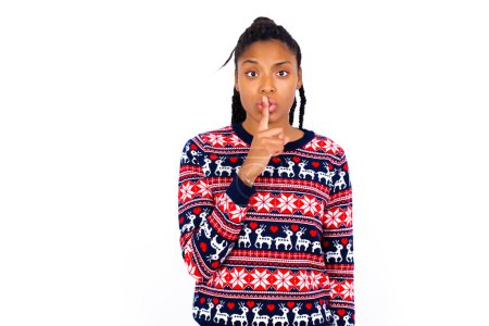 Téléchargez les photos : Jeune femme afro-américaine portant chandail de Noël ovre mur blanc fait un geste de silence, demande être calme. Ne dites pas mon secret ou ne parlez pas trop fort, s'il vous plaît.! - en image libre de droit