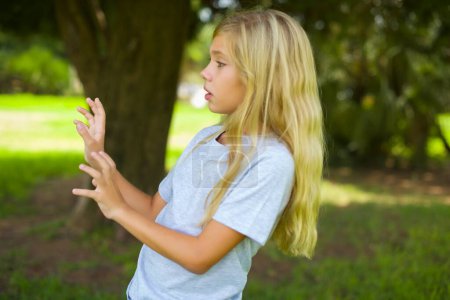 Photo for Displeased caucasian little kid girl wearing white t-shirt standing outdoor in the park keeps hands towards empty space and asks not come closer sees something unpleasant - Royalty Free Image