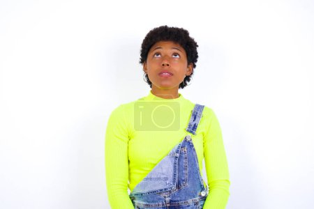 Téléchargez les photos : Jeune femme afro-américaine avec les cheveux courts portant le denim dans l'ensemble contre le mur blanc levant les yeux comme il voit quelque chose d'étrange. - en image libre de droit