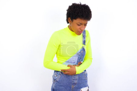Foto de Joven mujer afroamericana con el pelo corto con denim en general contra la pared blanca que sufre de dolor de estómago fuerte. - Imagen libre de derechos