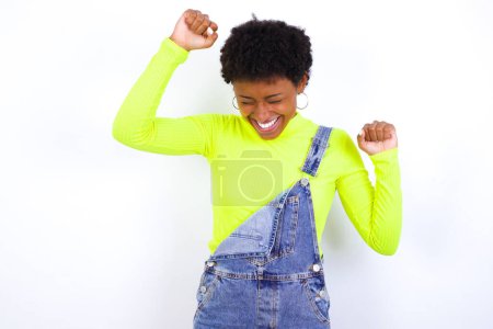 Foto de Atractiva joven afroamericana con el pelo corto vistiendo denim en general contra la pared blanca celebrando una victoria golpeando el aire con sus puños y una sonrisa radiante. - Imagen libre de derechos