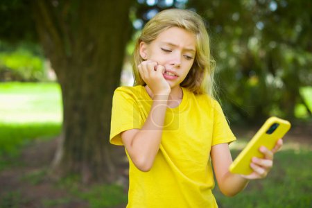 Téléchargez les photos : Portrait d'une petite fille caucasienne assez effrayée portant un t-shirt jaune debout à l'extérieur bavardant des ongles mordants après avoir lu des nouvelles effrayantes sur son smartphone. - en image libre de droit