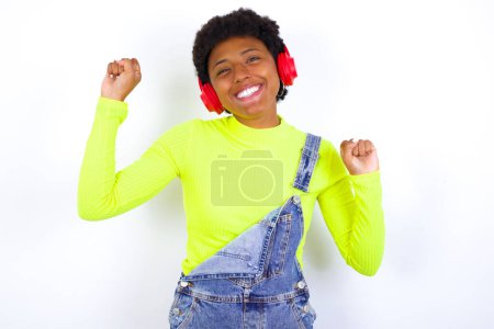 Téléchargez les photos : Jeune femme afro-américaine insouciante aux cheveux courts portant globalement du denim contre un mur blanc avec sourire aux dents soulève bras danses mouvements insouciants avec rythme de la musique écoute de la musique de la playlist via un casque - en image libre de droit