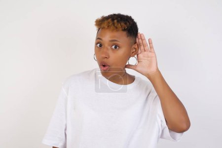 Oh my God! Funny astonished african-american woman wearing white t-shirt opening mouth widely, raising eyebrows and popping eyes out in surprise with hand near ear trying to listen to gossips.