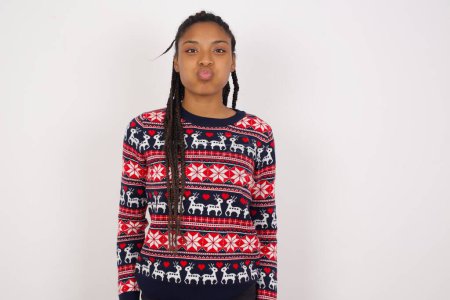 Téléchargez les photos : Femme afro-américaine portant un pull de Noël contre un mur blanc gonflant les joues avec un visage drôle. Bouche gonflée avec de l'air, expression folle. - en image libre de droit