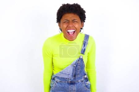 Photo for Stressful young African American woman with short hair wearing denim overall against white wall screams in panic, closes eyes in terror, keeps hands on head, finds out terrified news, can't believe it. - Royalty Free Image