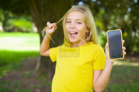 Foto de Foto de bonita niña caucásica usando camiseta amarilla de pie al aire libre demostrar la pantalla del teléfono mantenga las colas de pelo - Imagen libre de derechos