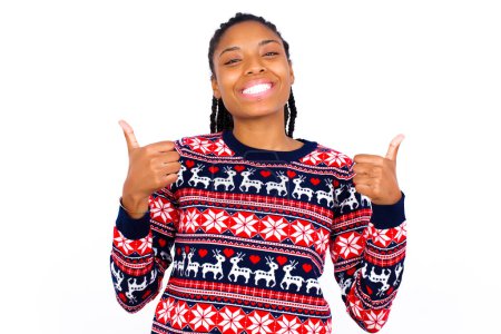 Téléchargez les photos : Femme afro-américaine optimiste portant un pull de Noël contre un mur blanc montrant les pouces levés avec des émotions positives. Concept de qualité et de recommandation. - en image libre de droit