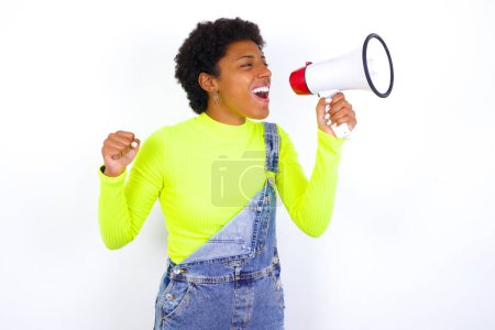 Foto de Joven mujer afroamericana con el pelo corto con denim en general contra blanco se comunica gritando fuerte sosteniendo un megáfono, expresando el éxito y el concepto positivo, idea de marketing o ventas. - Imagen libre de derechos
