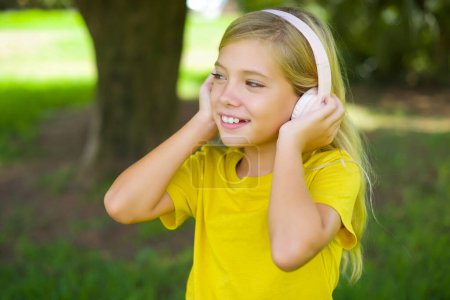 Photo for Caucasian little girl wearing yellow t-shirt standing outdoors wears stereo headphones listens music concentrated aside. People hobby lifestyle concept - Royalty Free Image