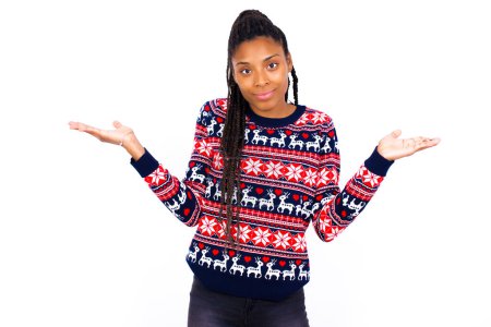 Photo for Puzzled and clueless African American woman wearing Christmas sweater against white wall with arms out, shrugging shoulders, saying: who cares, so what, I don't know. Negative human emotions. - Royalty Free Image