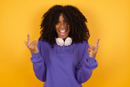 Foto de Retrato de joven mujer afroamericana expresiva con auriculares sobre fondo amarillo mostrando gestos rocosos - Imagen libre de derechos