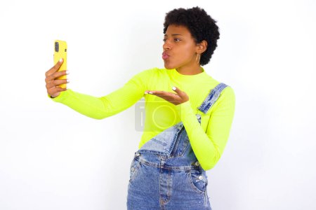 Foto de Joven mujer afroamericana con el pelo corto con denim en general contra golpes blancos beso de aire en la cámara del teléfono inteligente y toma selfie, envía mwah a través de llamada en línea. - Imagen libre de derechos